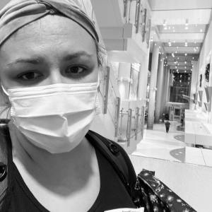 Kristen wearing a surgical mask, her eyes the main thing visible on her face. Her eyes look tired, her soul looks heavy with grief and purpose. Behind her is the entrance hallway to Riley Children's Hospital in Indianapolis. 